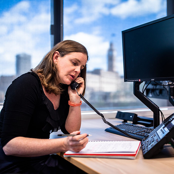 Office worker seated at a work desk, talking on the phone and holding a pen, ready to take notes in an A4 sized notepad.